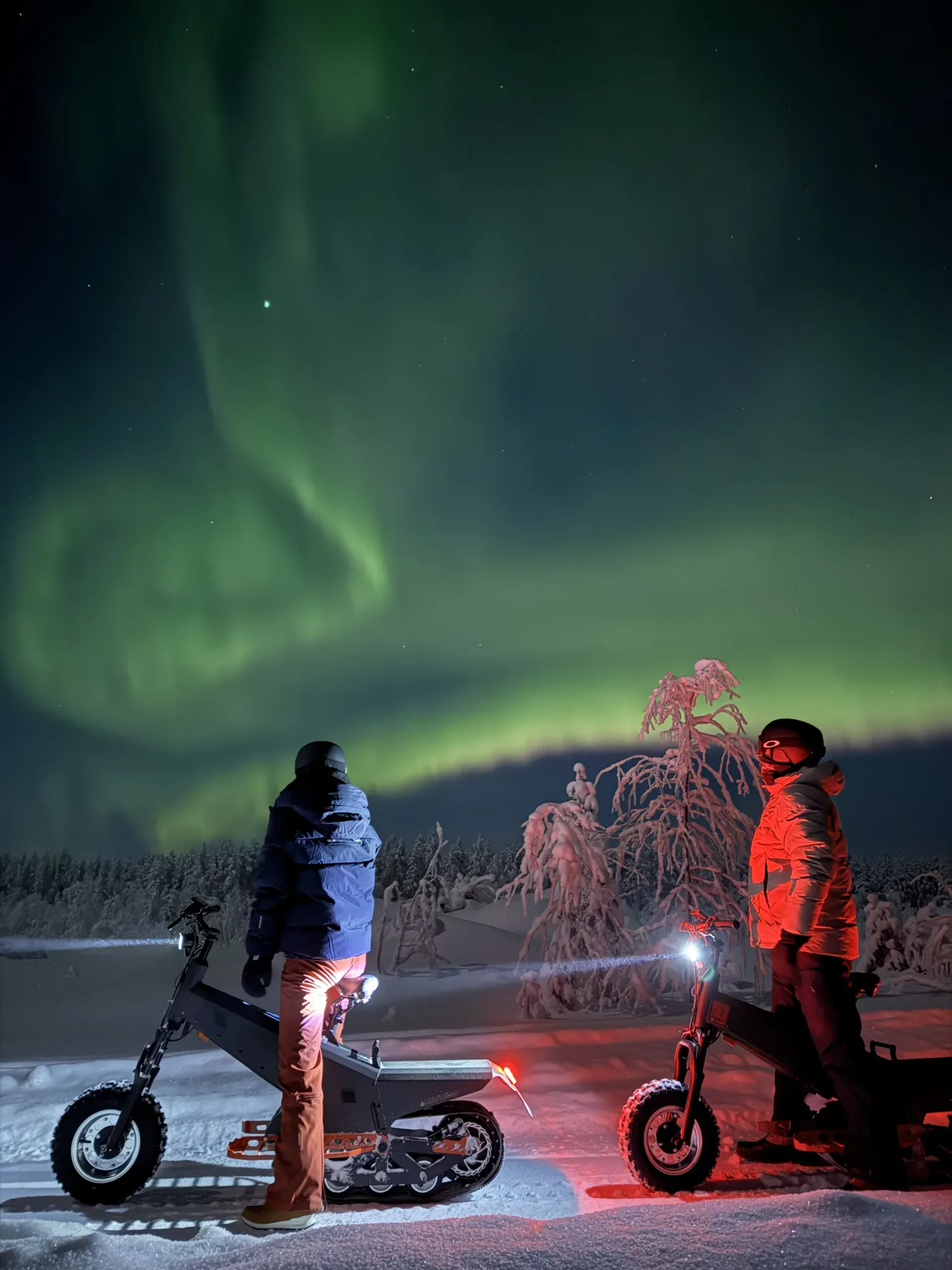 Two people standing in the snow at night beside a frost-covered tree lit red by a headlamp, looking up at green aurora spreading across the sky